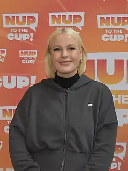Purcell standing behind a "Nup to the Cup" backdrop wearing a jacket and turtleneck