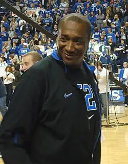 A man, wearing a black jacket and black T-shirt, is walking onto the basketball court.