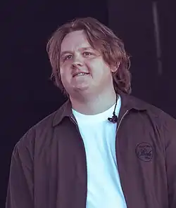 A man with shoulder-length brunette hair wearing a brown jacket with a plain white t-shirt. An earpiece is visible on his shirt.