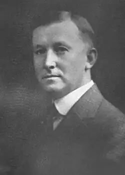 A young man with dark hair wearing a white shirt, dark tie, and dark jacket