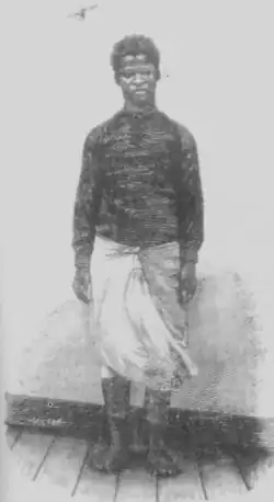A young boy in a dark top and a white garment tied around his waist standing barefoot in front of a blank wall.