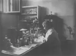 Photograph of Marie Goldsmith, wearing a lab coat, working at a laboratory desk covered with papers and bottles