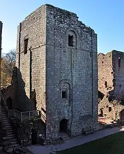 A square stone keep dominates the picture, sat behind a patch of green grass; the keep has a doorway at ground level, with two windows irregularly placed above it.