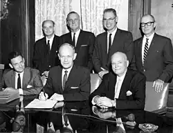 Governor Farris seated front and center, with pen and paper, in a group of 7 men.