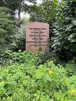 Pink memorial stone amid lush greenery, in the foreground there are light green leaves with scattered yellow flowers with four petals. On the memorial there are six names. Prof. Friedrich Fedde 1873–1942; Katharina Fedde 1879–1950; Friedrich Karl Fedde 1904–1946; Gertrud Fedde 1909–1979; Dr. Wolfdiether Fedde-Woymode 1912–1987; Ilse Fedde-Woymode 1914–(Obscured)