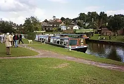 A narrow boat moored alongside grassy area. Houses can be seen in the background.