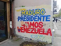 A blue, red and yellow graffiti on a wall reading "MADURO PRESIDENTE / VAMOS VENEZUELA"