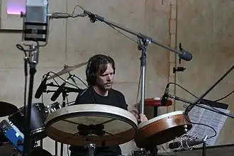 Greg Ellis wearing a black t-shirt, sitting behind a modified drum kit and appearing to play two hand drums