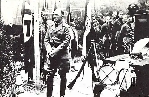 A jacketed Nazi of rank turned away from men at attention