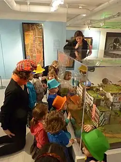 Young children in hats looking at a glass showcase containing the model of a building