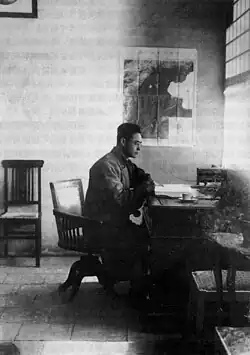 A black and white photo of Gu Jiegang seated at a desk in an office. A map of the area around Beijing is visible