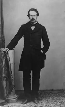 A black and white, full-length portrait of an middle-aged bearded man wearing a dark suit, a bow tie, and resting his right hand over a small column.