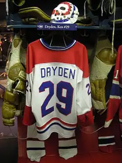 White hockey jersey with red and blue numbers and trim hanging from a shelf with a goalkeeper mask and padded equipment