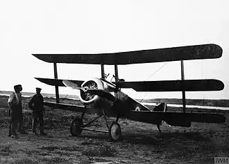 Front three-quarter view of military triplane on landing ground, with two men standing beside it