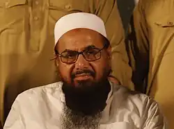 A color photograph of a seated, bearded, bespectacled man wearing an Islamic skullcap