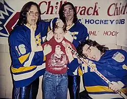 Three bespectacled brothers in blue hockey uniforms surround a child in a red shirt and jeans, who is a fan of the trio.