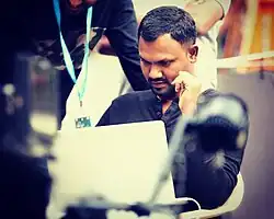 Harish Komme sitting and looking down at a laptop, with left elbow propped up on armrest; two people are partly visible in background, one wearing a lanyard