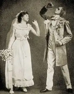 Young white bride and groom standing in Victorian wedding clothing; she holds a bouquet and gazes at him, as he strikes a melodramatic pose