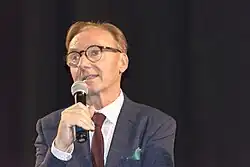 close-up of Ondřej Havelka wearing a pattered white shirt, burgundy tie, grey/blue suit with green pocket square, speaking into a microphone in front of a dark backdrop