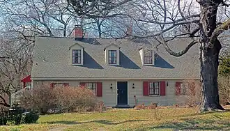 A light yellow one-and-a-half-story wooden house seen from its front, with a large bare tree obstructing part of the view at upper right. It has reddish-orange shutters on the windows and three dormer windows in its shingled roof.