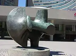 Photograph of a large bronze abstract sculpture, in front of a glass and concrete building.