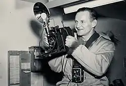 An image of Henry Busse in 1947. He's smiling and holding a flash bulb camera.