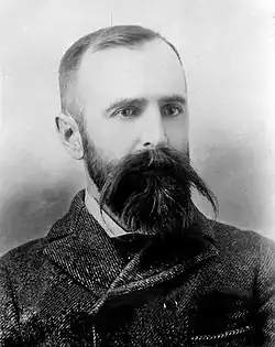 Portrait shot of a man perhaps around 40 (Wilson). He is dressed in archaic clothing, with a checked suit jacket close around his neck. His hair is cut close, but he wears a beard and an impressive mustache, with straight hair from it extending well to each side of the face.