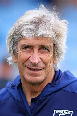 A photograph of a gentleman in his 60s. He is sitting on a red side bench. He is wearing a navy blue hooded jacket with a Manchester City crest on the left breast and a white logo of the Nike sponsor on the right breast.
