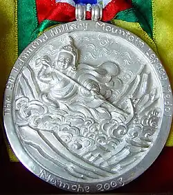 A silver medallion with an embossed image of Guru Rinpoche using a hand-drawing plow to harrow the sacred Rolwaling Valley. Around the upper edge is the text "Sir Edmund Hillary Mountain Legacy Medal"; at bottom is the text "Namche 2003"