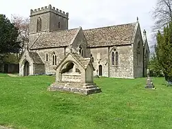 Gray stone building with small square tower at left hand end. In the foreground is grass with a small tiled memorial.