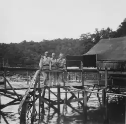 Black-and-white photo of three men in tropical military uniform. They are standing outdoors on an elevated walkway above a body of water in a tropical jungle locale. A hut-style building is visible to the right.