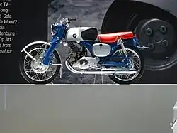 A pristine-looking 1960s-era single-cylinder motorcycle with a blue frame and bright red seat, and a Honda wing logo on the gas tank.