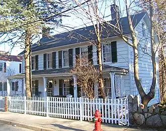 A white wooden house with full front porch, black window shutters and chimneys at either end behind a white picket fence and some bare trees, lit by sunlight from the right
