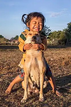 A young girl smiling broadly, standing behind a small tan dog and hugging it