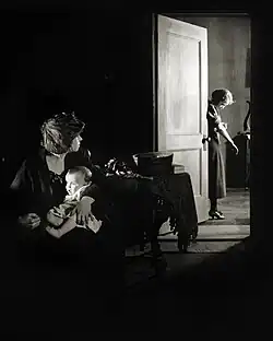 Shadowed interior with a woman cradling a baby in the foreground while another woman in the doorway injects morphine into her arm