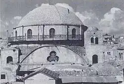 A large domed roof sits upon a bricked parapet, which includes arched windows, three of which are visible. The base of the parapet is surrounded by a narrow veranda, which is fenced with decorative metal grating. Below this, a central clover shaped window lies at the top of an arched wall. Two supporting towers stand on the left and right, each containing an arched window. The right tower, capped with a small dome, is a storey higher, reaching to the top of the bricked parapet, which supports the domed roof.