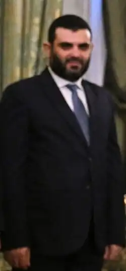 Bearded man wearing a black suit with a blue tie. His beard is black.