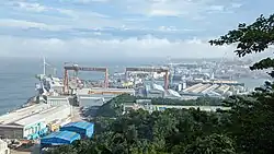 A photo overlooking Ulsan Shipyard. Enormous red gantry cranes stand beside warehouses, and wind turbines can be seen in the distance.