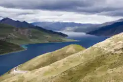 Yamdrok Tso, a large high-altitude lake in Tibet, surrounded by rolling green hills and distant mountains under a cloudy sky.