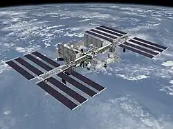 A planform view of the International Space Station is backdropped by the limb of the Earth. In view are the station's four large, blue-colored solar array wings, two on either side of the station, mounted to a central truss structure. Further along the truss are six large, white radiators, three next to each pair of arrays. In between the solar arrays and radiators is a cluster of pressurised modules arranged in an elongated T shape, also attached to the truss.