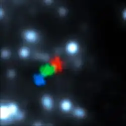 A blurry image of black space illuminated by white blobby stars. Three different observations of a gas cloud from different years can be seen, showing the cloud moving and deforming in shape.
