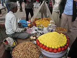 A street food vendor spoons onions onto newsprint to serve. His stall has several ingredients, including puffed rice, peanuts, tomatoes, lemons, and chillis.