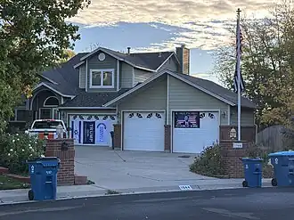 This photograph depicts the suburban house at 1944 Quail Circle in Lafayette, Colorado, commonly identified as the home base of the International Slide Rule Museum, with a three-bay garage, brick pillars at the driveway, curb address marker “1944,” and recycling carts along the street; banners are hung on the left garage, and a flagpole stands at right under a patterned morning sky.