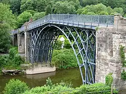An ornate bridge made of iron, passing over a small, lush valley