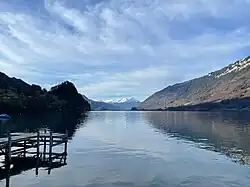 Panoramic view of a tranquil Swiss lake surrounded by mountains, with a wooden dock in the foreground and snow-capped peaks under a partly cloudy sky in the distance.