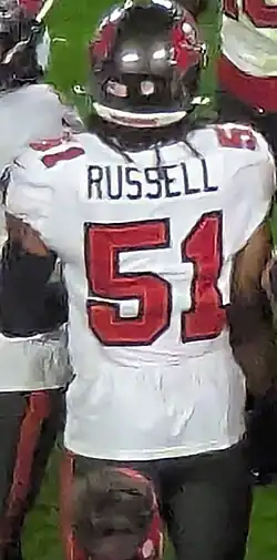 J.J. Russell standing on the sideline wearing a jersey representing his team, the Tampa Bay Buccaneers, with "Russell" written in black at the top of the rear of his jersey and "51", the number he wears, in red below his name. He is also wearing a gray Buccaneers helmet.