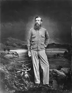 A black and white portrait of a bearded man. The man wears a coat and pants, and is posing in front of a painting of a large lake.