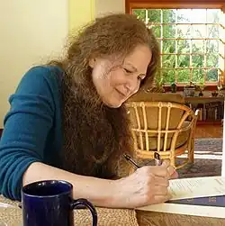 a photograph showing Jane Hirshield, a white woman with long curly hair, seated at a table and writing