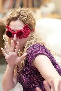 A woman with curly blonde hair wears oversized red crochet sunglasses and a patterned purple top while playfully holding her hands up toward the camera
