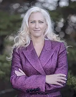 A woman with long wavy platinum blonde hair wearing a textured fuchsia blazer stands outdoors with her arms crossed